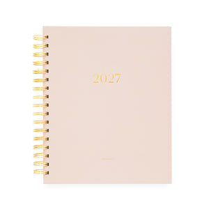 A 2027 spiral-bound planner with a pale blush pink cover and gold foil lettering, bound with a gold wire coil and '2027' and 'planner' foil stamped on the cover