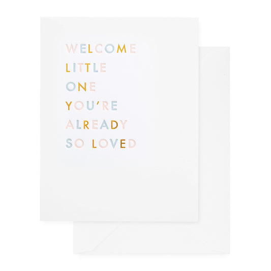 A greeting card with the text 'WELCOME LITTLE ONE YOU'RE ALREADY SO LOVED' printed on it in pink, blue and gold foil paired with a white envelope