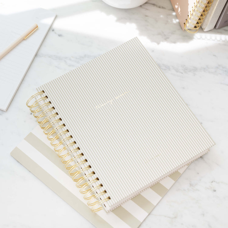 A flax-colored spiral-bound planner with thin vertical stripes on its cover, styled on a white marble desk alongside other stationery items.