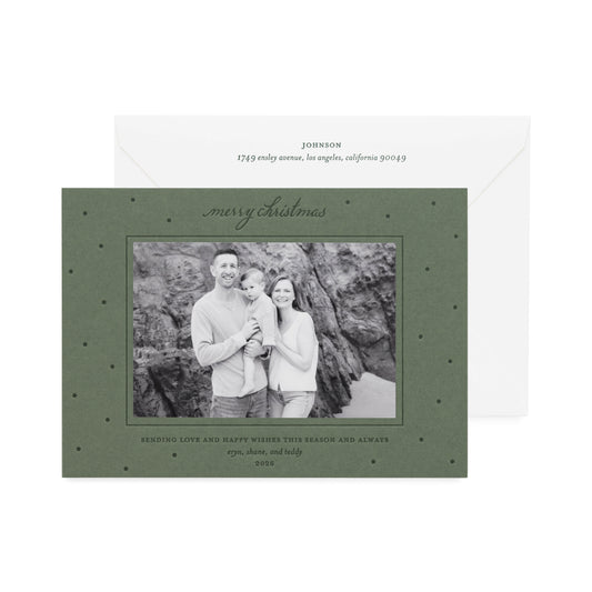A Christmas holiday photo card on moss green paper and a central black-and-white photo of a family, shown with a white envelope behind it with dark green text. The card is printed with 'merry christmas sending love and happy wishes this season and always'