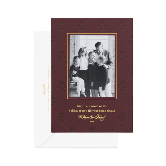 A holiday photo card with a family photo on mulberry paper, featuring gold foil printed with 'may the warmth of the holiday season fill your home always' and mulberry letterpress floral details, with a white envelope.