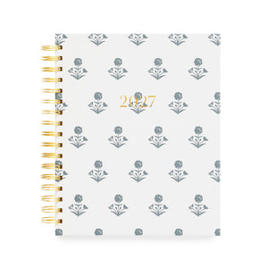 A 2027 spiral-bound planner with a white cover with a blue floral pattern and gold foil text reading '2027' in the center. The left side features a gold-tone spiral coil binding.