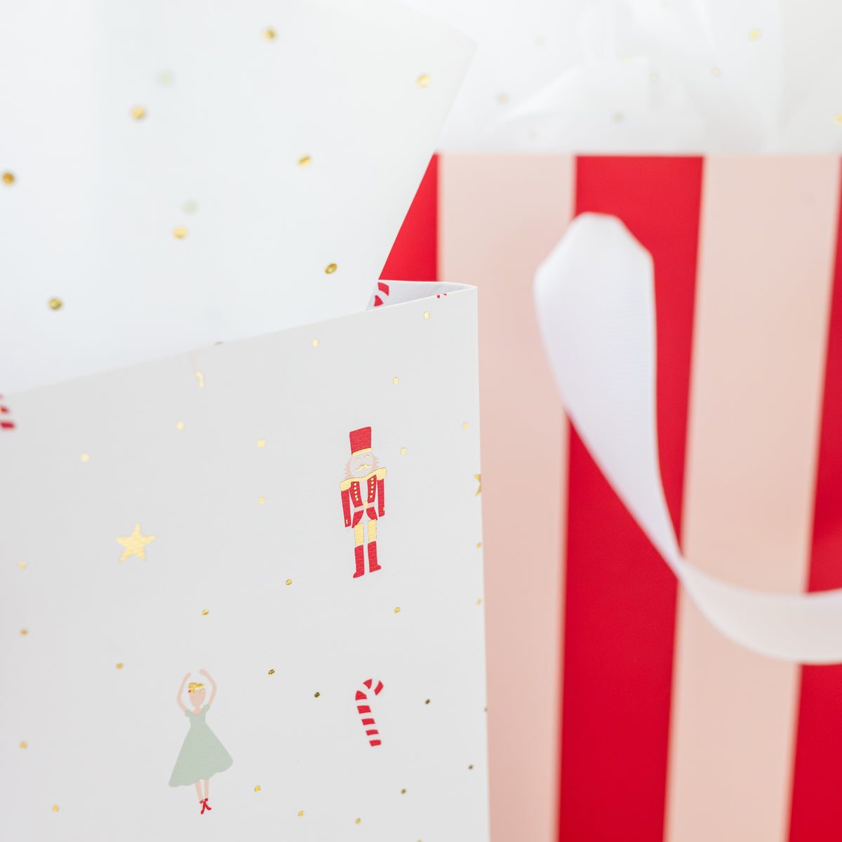 A close-up of a white gift bag decorated with gold foil dots and stars, a holiday nutcracker pattern, alongside a red and pink striped gift bag with white ribbon handle