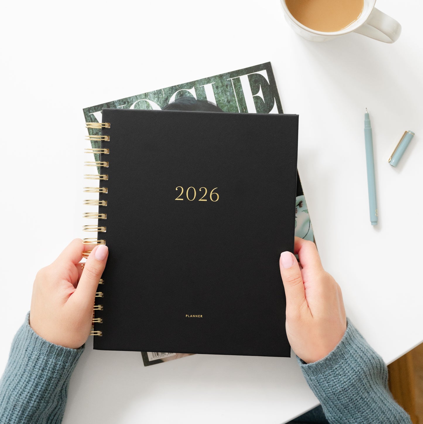 Person holding a black planner with '2026' on it, placed on a white surface with a cup of coffee and magazine