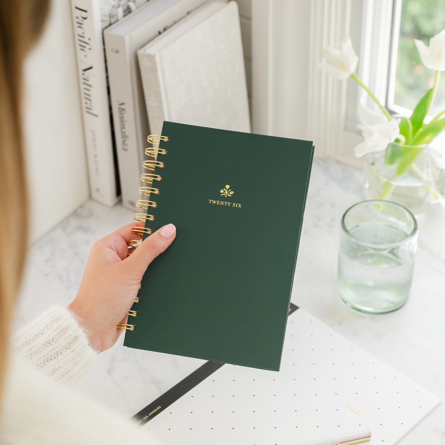 Hand holding a green spiral planner with 'Twenty Six' in gold foil on a desk with books and a glass of water.