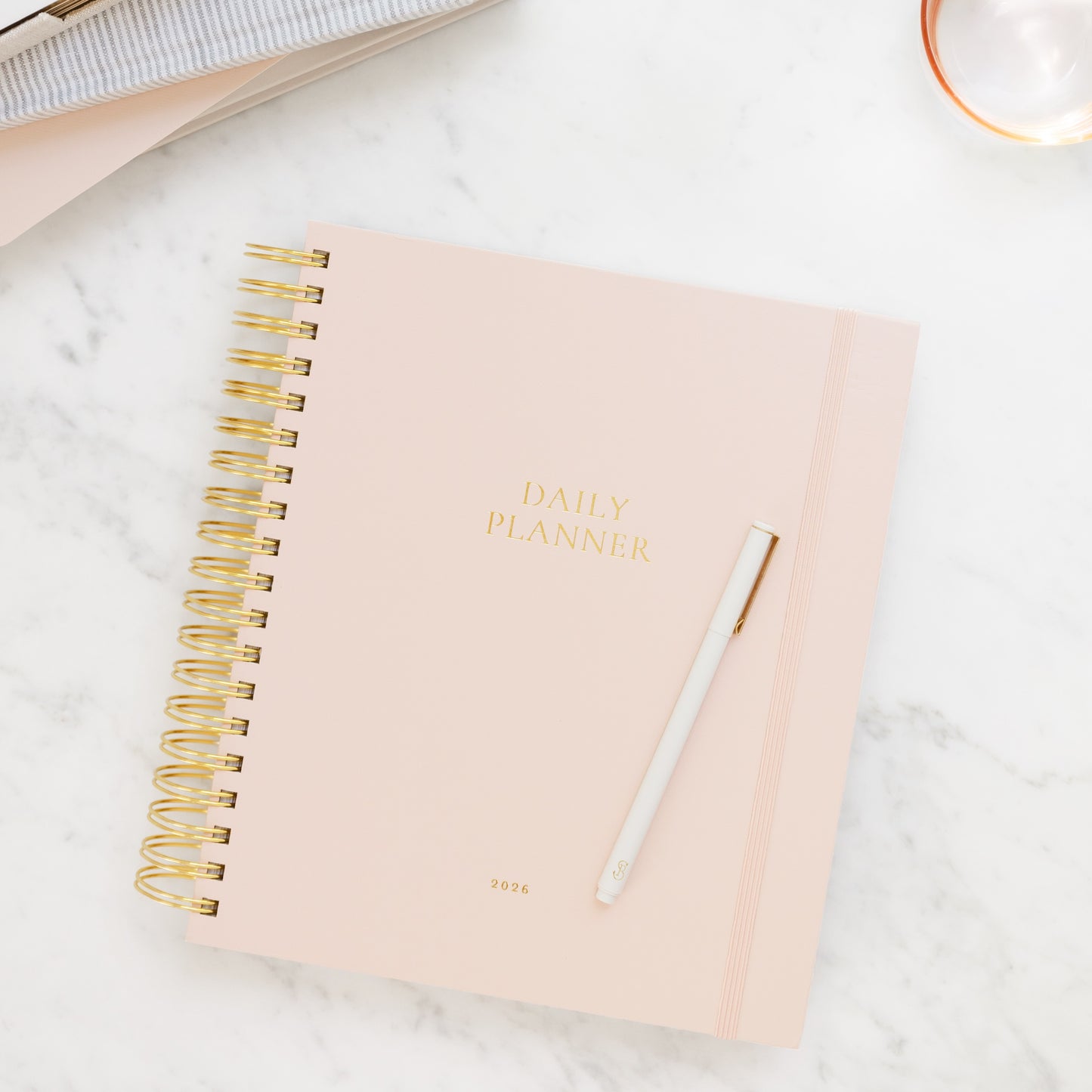 Blush pink daily spiral planner with gold spiral binding on a marble surface with journals in background