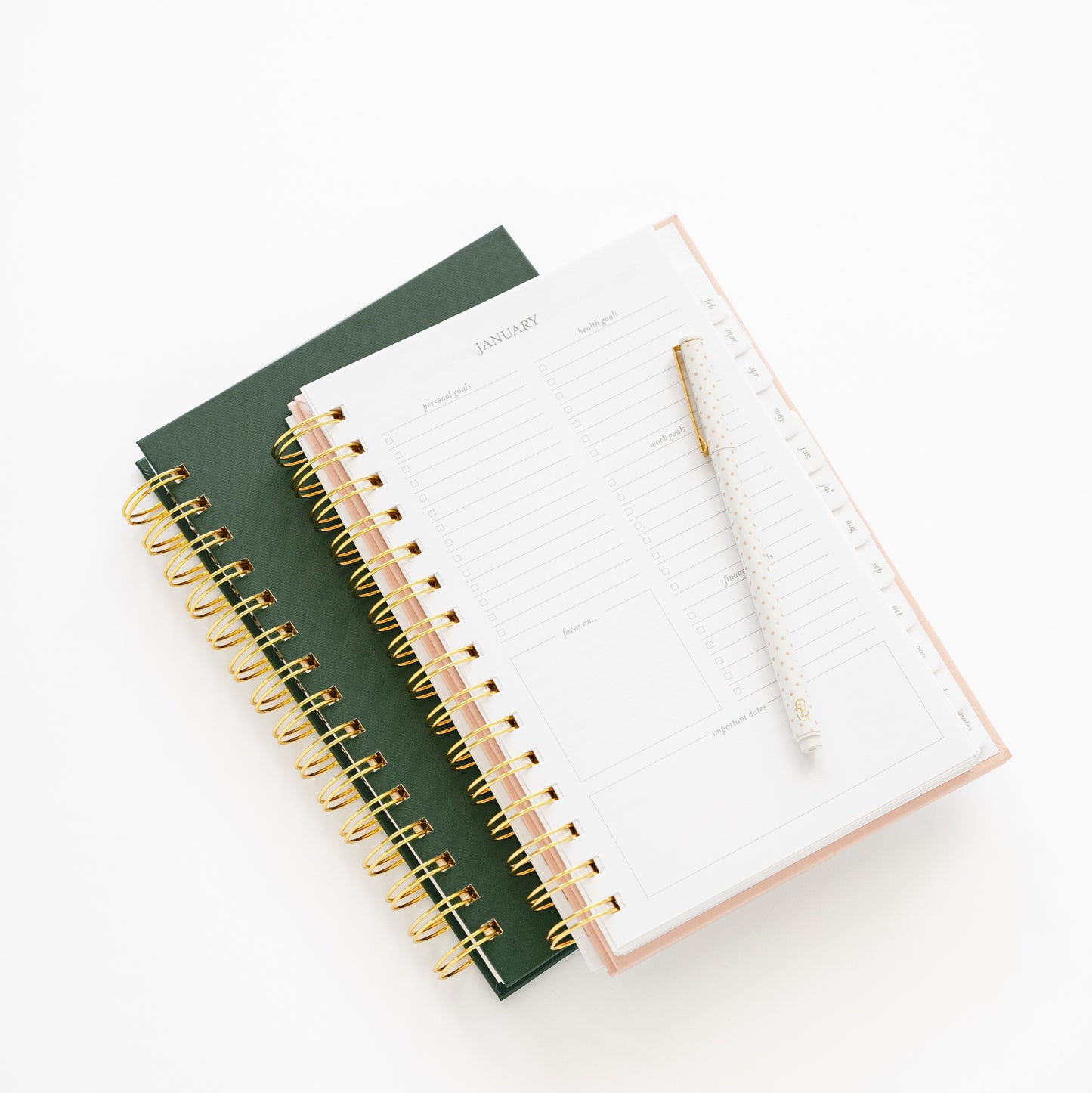 Two small spiral-bound planners, one green and one blush open to monthly goal page with gold spirals