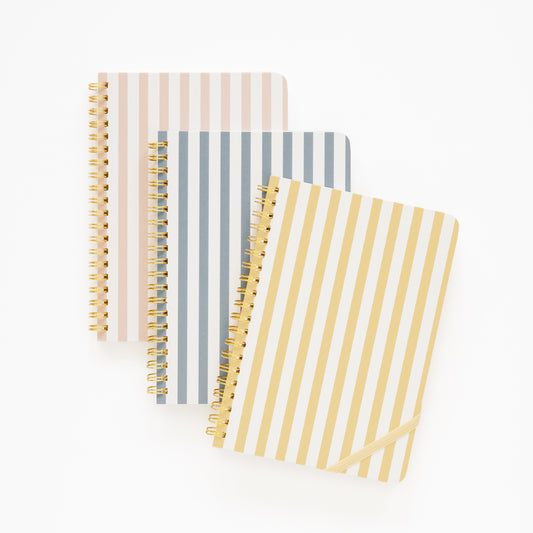 Three spiral notebooks with striped covers in beige, blue, and yellow with gold spiral binding