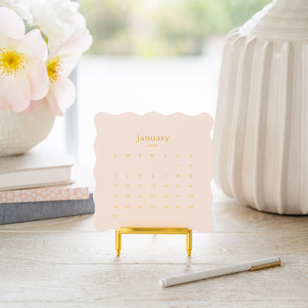 A small January 2027 desk calendar with a scalloped pale pink border and gold foil lettering, displayed on a gold stand on a tabletop. A stack of journals, light, and flowers in background