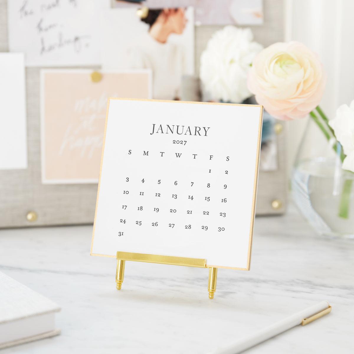 A small January 2027 monthly desk calendar stand on a gold metal easel, displayed on a marble surface with flowers and an inspiration board in background