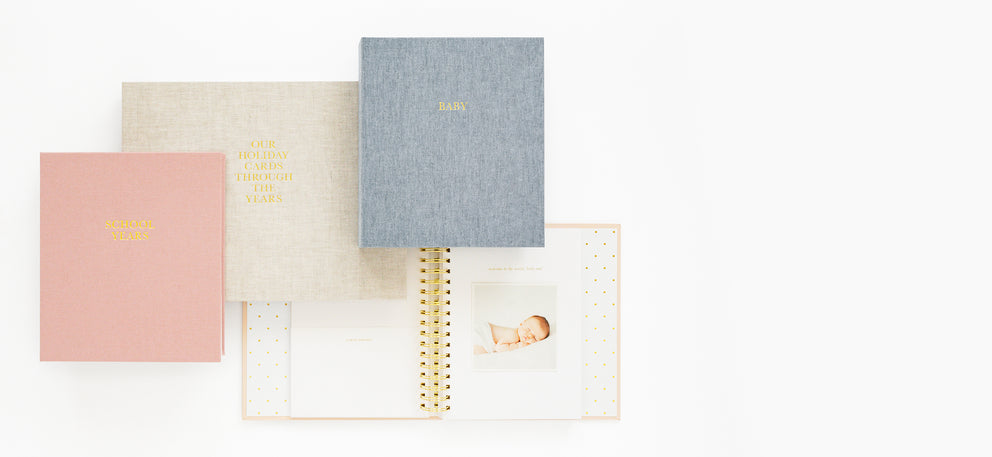 Rose pink school years book, flax book with our holiday cards through the years, and blue fabric book with baby on cover with a pink baby book open to a photo of a baby on the page 'welcome to the world little one'