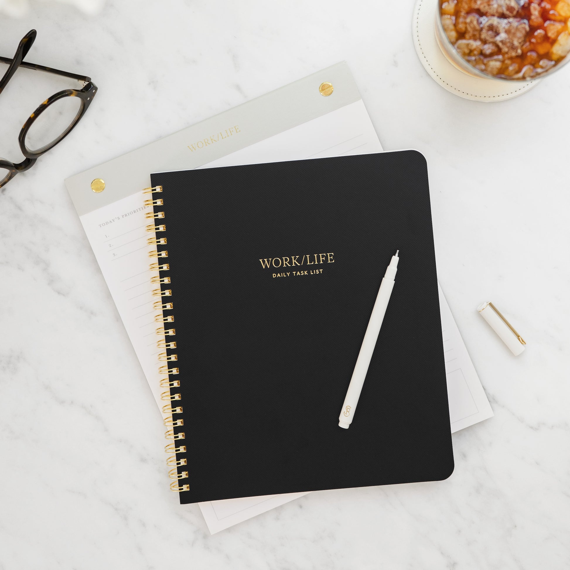 Black spiral bound notebook with 'Work/Life' daily task list on the cover on a marble surface with glasses and coffee.