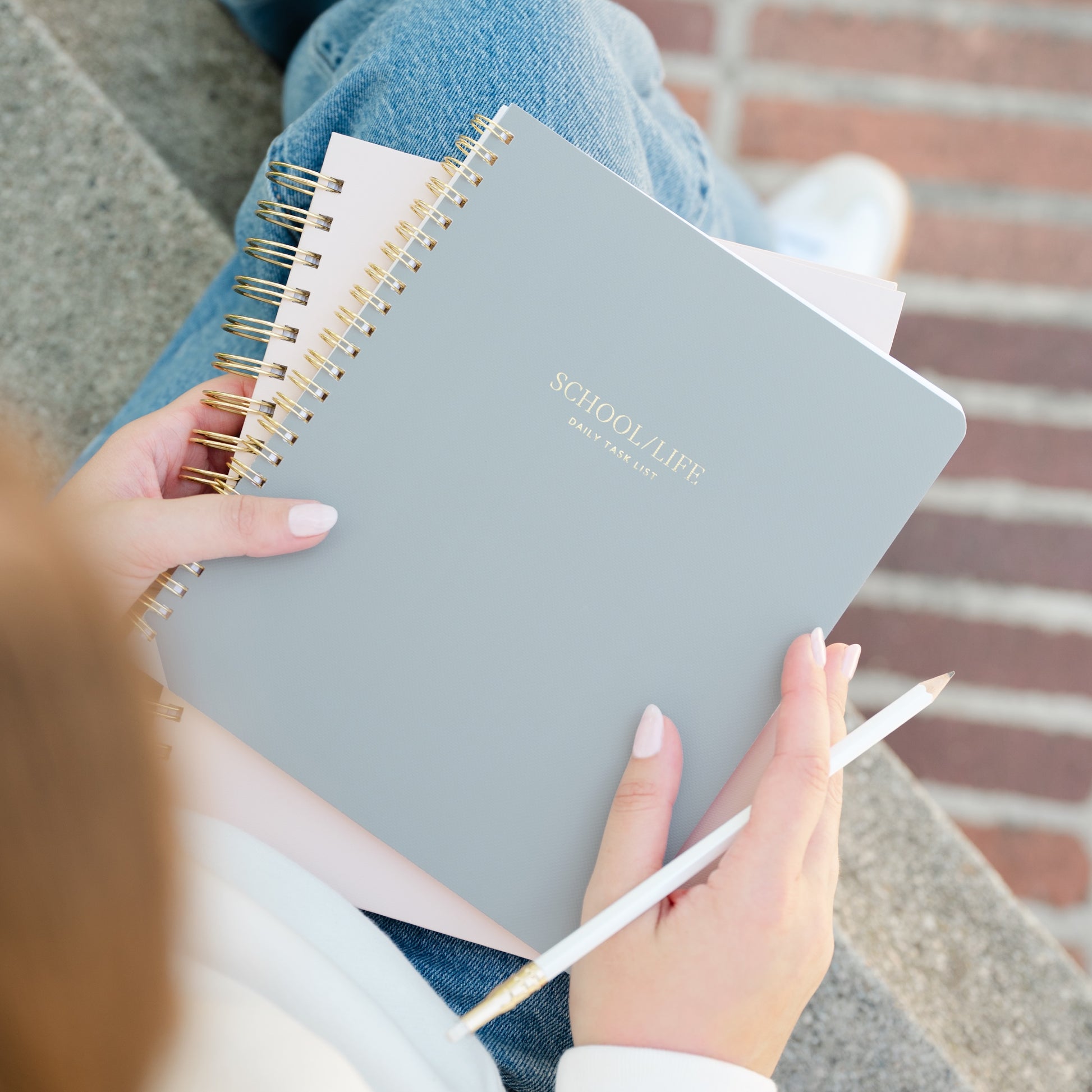 Person holding a light blue spiral bound notebook with 'SCHOOL/LIFE' daily task list in gold foil on cover, sitting on a bench.