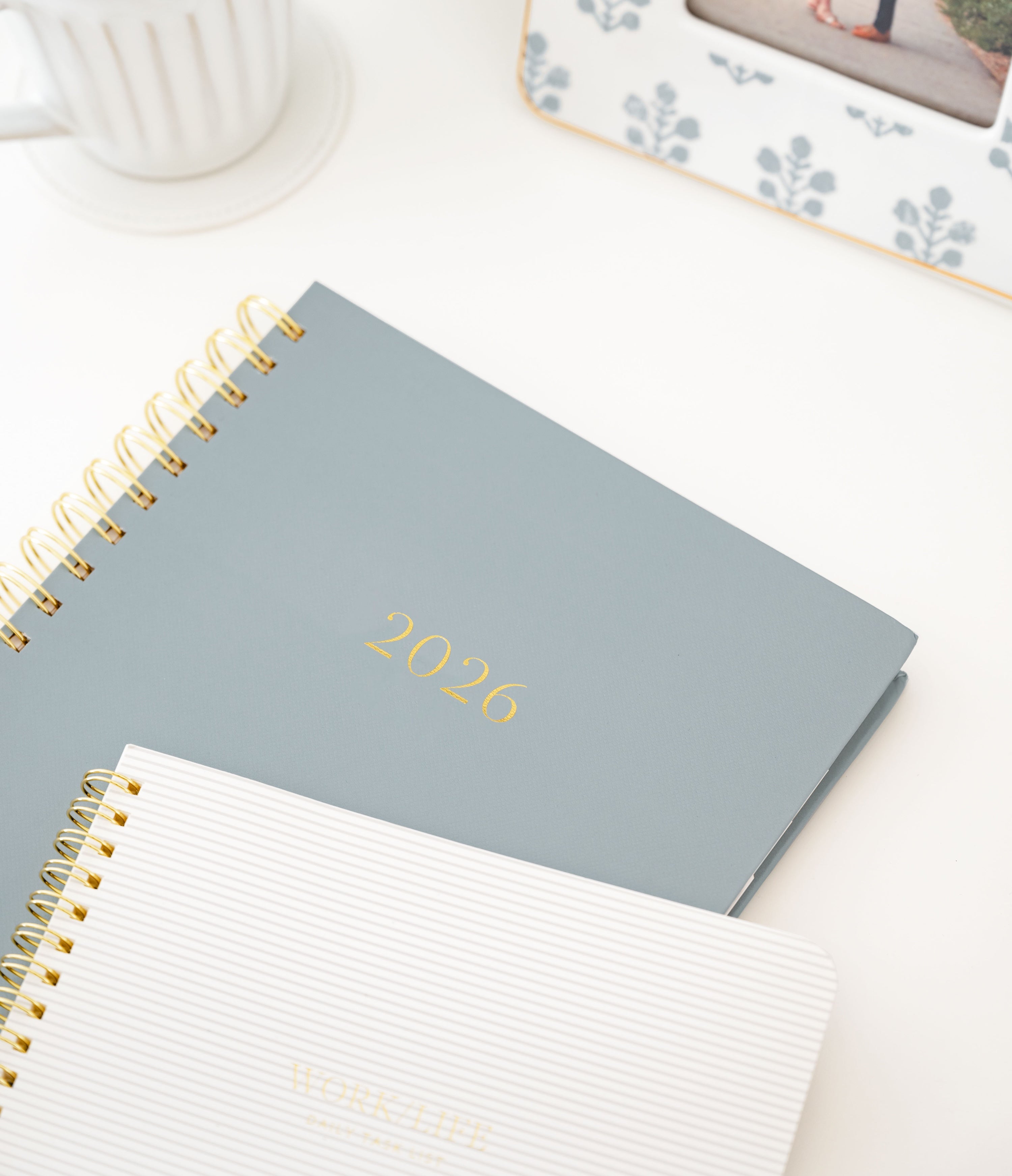 Spiral-bound dusty blue planner with '2026' in gold foil paired with a grey stripe notebook