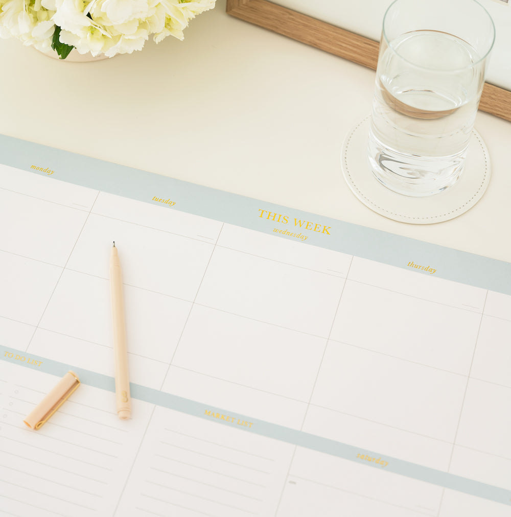 Oversized blue and white weekly desk notepad on desk with a pale pink pen and glass of water