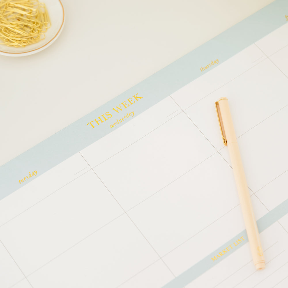 Oversized blue and white weekly desk notepad with a pale pink pen and close up on 'this week' gold foil print