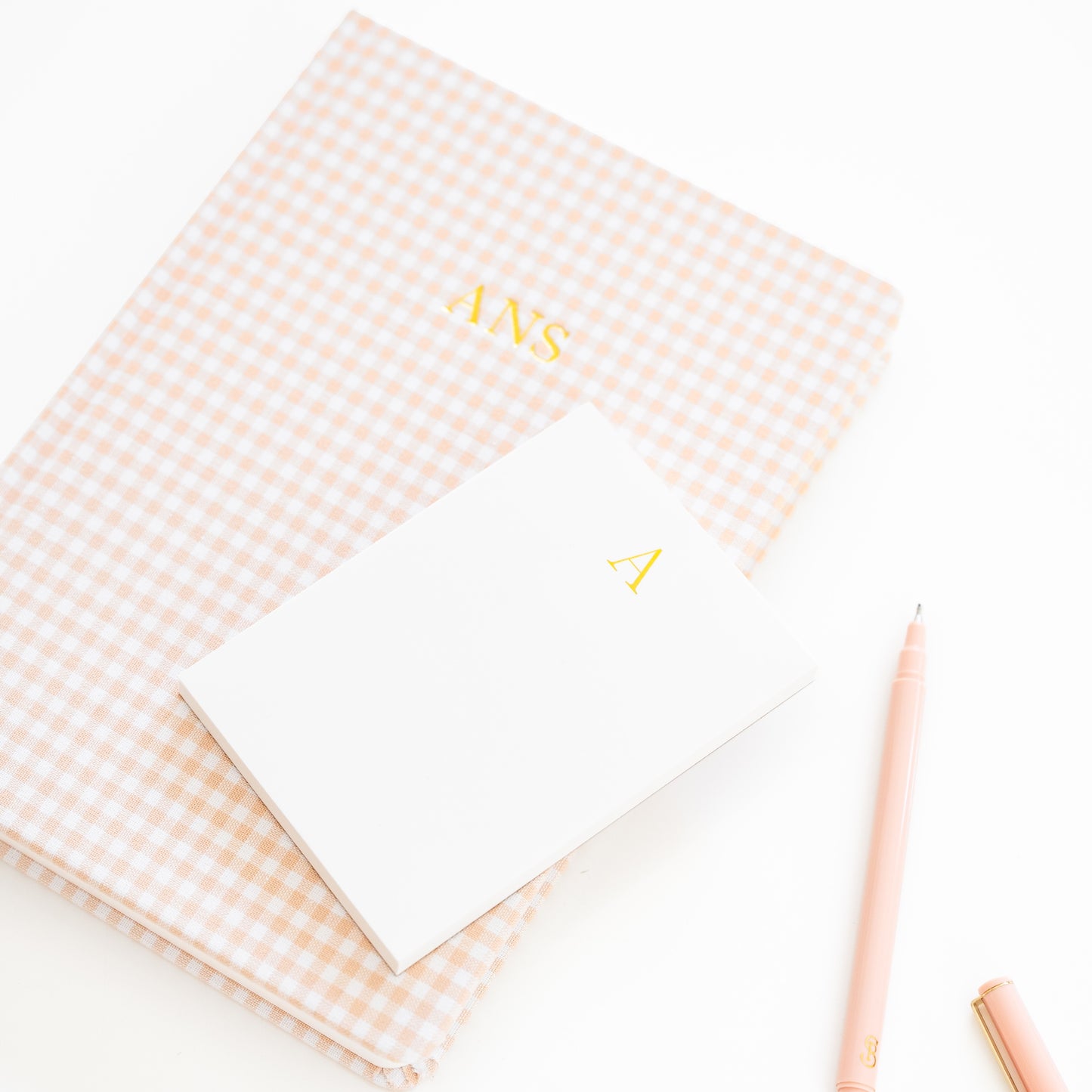 Pink gingham fabric journal with 'ANS' on cover paired with a white mini notepad with 'a' in gold foil and a pink pen