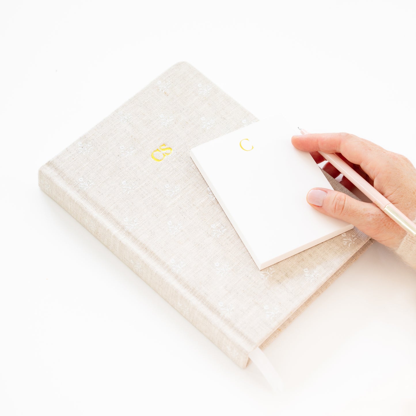 Hand holding a white notepad with gold 'c' on top of a flax journal with 'cs' lettering in gold foil