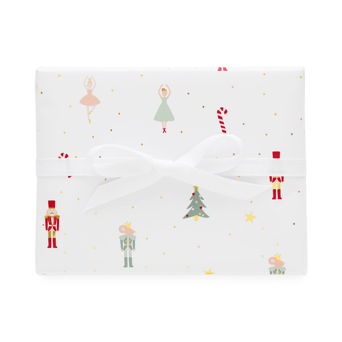 A square gift box wrapped in white wrapping paper with a festive Christmas print featuring nutcrackers, ballerinas, candy canes, a Christmas tree, and gold dots, tied with a white ribbon