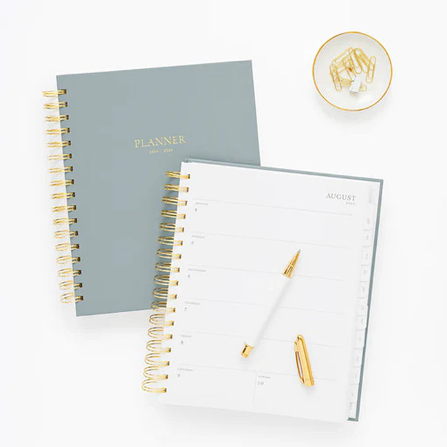 Two academic planners, one open and one on the cover, with a white and gold pen on a white desk