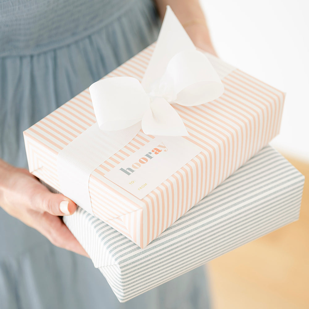Stack of two wrapped gifts in hands in blue stripe and pink stripe patterns tied with a big white grosgrain bow and a hooray sticker with to and from sections