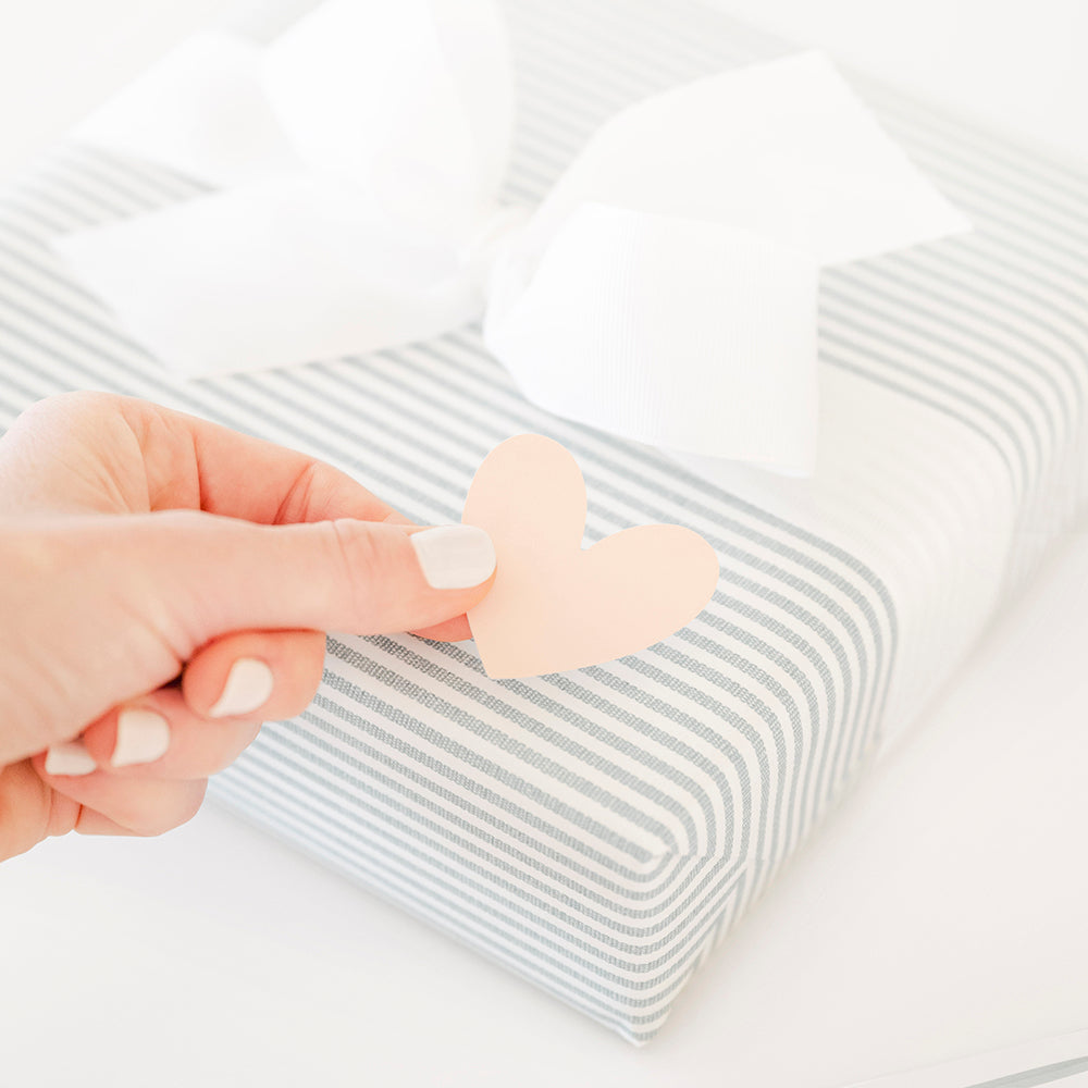 Close up of a blue and white striped wrapped gift with a white bow and someone putting on a pink heart sticker