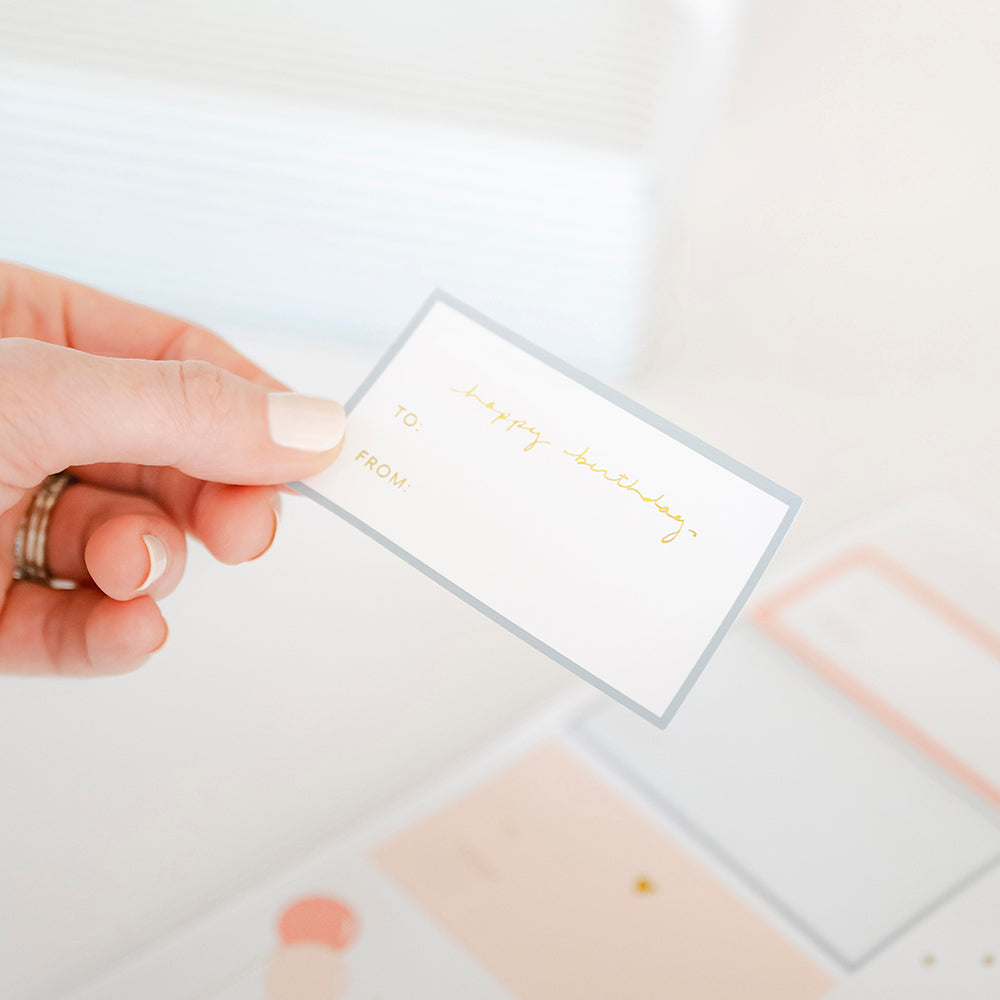 Hand holding birthday sticker "happy birthday to and from" with a blue border over a blurred sticker sheet of multiple birthday stickers