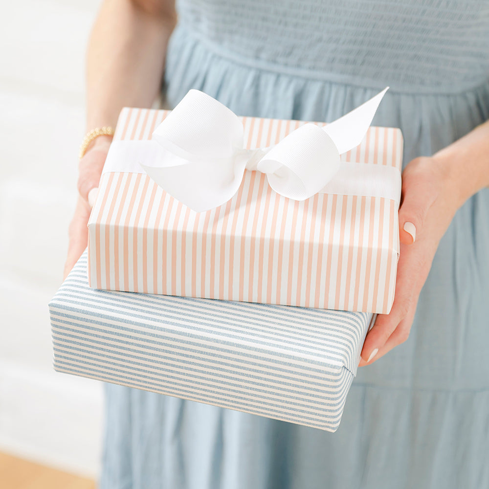 Stack of two wrapped gifts in hands in blue stripe and pink stripe patterns tied with a big white grosgrain bow