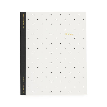A minimalist 2027 monthly planner with a cream-colored cover decorated with a grid of small black dots and gold foil text with '2027' with a black tape binding