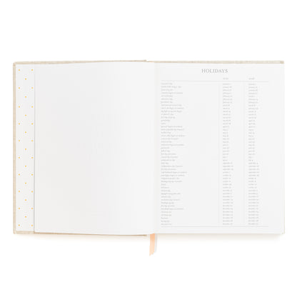 An open hardcover planner showing a printed holiday schedule page for 2027 and 2028, with blank lined pages on the left and a pink and white ribbon markers