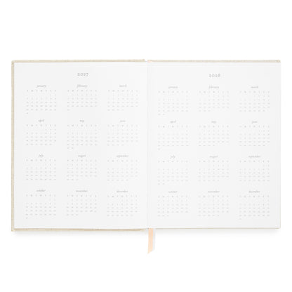 An open planner showing two blank interior spread with 2027 and 2028 calendar grids and a pink and white ribbon marker