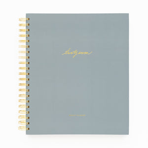 A 2027 spiral-bound planner with a matte slate blue cover and gold foil lettering with 'twenty-seven' and 'weekly planner' stamped on cover with a gold coil spiral