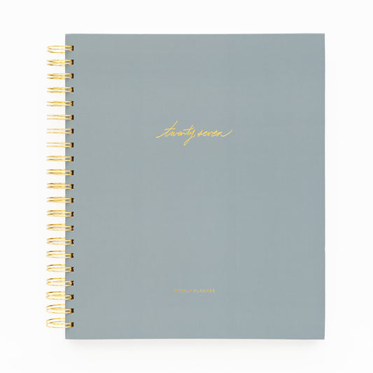 A 2027 spiral-bound planner with a matte slate blue cover and gold foil lettering with 'twenty-seven' and 'weekly planner' stamped on cover with a gold coil spiral