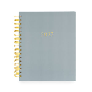A 2027 spiral-bound planner with a matte slate blue cover and gold foil lettering, bound with a gold wire coil