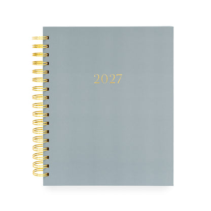 A 2027 spiral-bound planner with a matte slate blue cover and gold foil lettering, bound with a gold wire coil