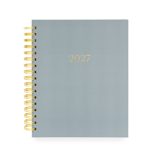 A 2027 spiral-bound planner with a matte slate blue cover and gold foil lettering, bound with a gold wire coil