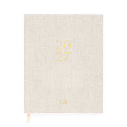 A hardcover linen planner in flax linen with '2027' in gold foil and pink and white ribbon markers