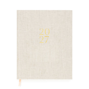 A flax linen fabric bound 2027 planner with gold foil '2027' on the cover and pink and white ribbon markers