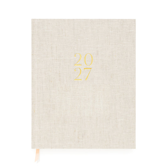 A flax linen fabric bound 2027 planner with gold foil '2027' on the cover and pink and white ribbon markers