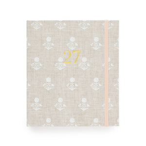 A 2027 fabric planner  with a beige linen cover, featuring a white floral pattern, a gold foil '27' on the front, and a pale pink elastic closure band