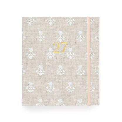 A 2027 fabric planner  with a beige linen cover, featuring a white floral pattern, a gold foil '27' on the front, and a pale pink elastic closure band