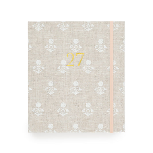A 2027 fabric planner  with a beige linen cover, featuring a white floral pattern, a gold foil '27' on the front, and a pale pink elastic closure band