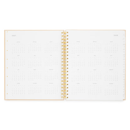 An open spiral-bound planner showing monthly calendar pages for 2027 and 2028, with a light pink cover and gold spiral binding