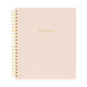 A blush pink spiral-bound planner with gold wire binding and gold foil text on the cover with 'twenty-seven' and 'weekly planner'