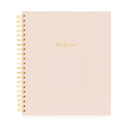 A blush pink spiral-bound planner with gold wire binding and gold foil text on the cover with 'twenty-seven' and 'weekly planner'
