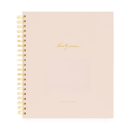 A blush pink spiral-bound planner with gold wire binding and gold foil text on the cover with 'twenty-seven' and 'weekly planner'