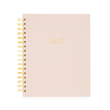 A 2027 spiral-bound planner with a pale blush pink cover and gold foil lettering, bound with a gold wire coil and '2027' and 'planner' foil stamped on the cover