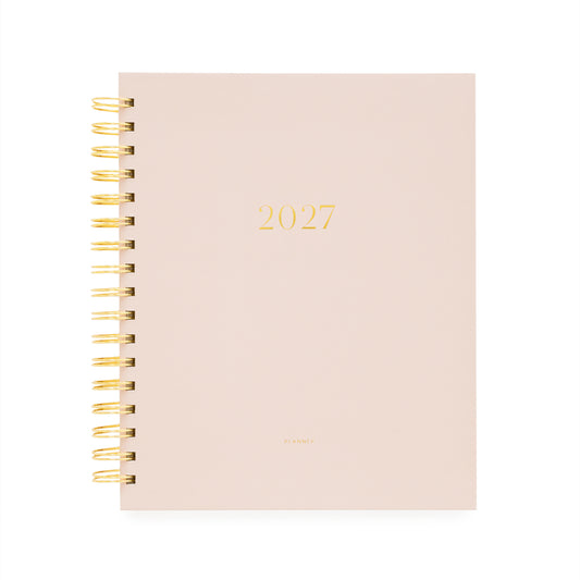 A 2027 spiral-bound planner with a pale blush pink cover and gold foil lettering, bound with a gold wire coil and '2027' and 'planner' foil stamped on the cover