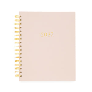 A 2027 spiral-bound planner with a pale blush pink cover and gold foil lettering, bound with a gold wire coil