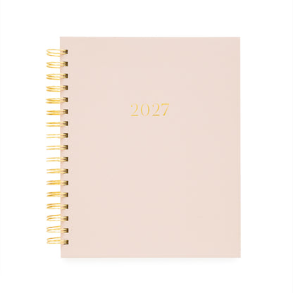 A 2027 spiral-bound planner with a pale blush pink cover and gold foil lettering, bound with a gold wire coil